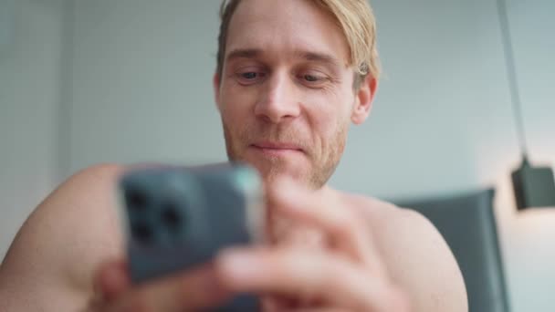 笑金发碧眼的运动员 赤身裸体 电话发短信 坐在床上朝旁边看 — 图库视频影像