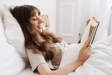White young woman smiling and reading book while lying in bed at home