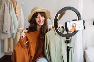 Beyaz kadın evde cep telefonundan selfie çekerken kıyafetlerini gösteriyor.