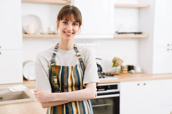 White young woman wearing apron smiling while standing in kitchen at home