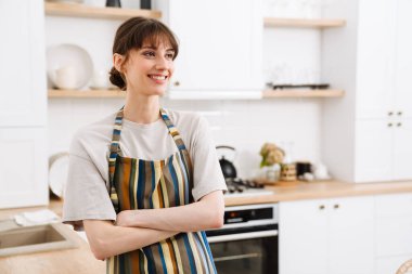 Evdeki mutfakta dururken önlük takan beyaz bir kadın gülümsüyor.