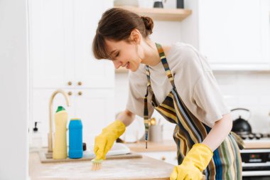 White young woman in gloves smiling while cleaning table with brush at home