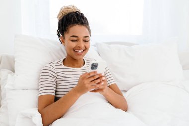 Genç siyahi kadın evde otururken gülümsüyor ve cep telefonu kullanıyor.