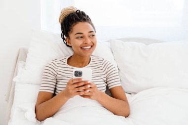 Genç siyahi kadın evde otururken gülümsüyor ve cep telefonu kullanıyor.