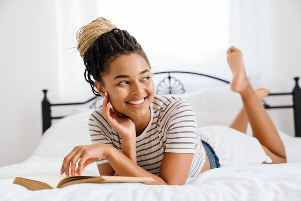 Young black woman reading book while lying on bed at home