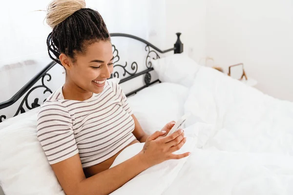 Young Black Woman Smiling Using Cellphone While Sitting Bed Home — Stock Fotó