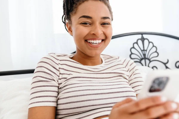 Genç siyahi kadın evde otururken gülümsüyor ve cep telefonu kullanıyor.