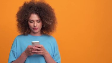 Smiling curly haired blonde woman wearing blue t-shirt typing by phone in the orange studio