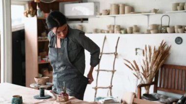 Gülümseyen kadın çömlekçi önlüğünü çıkarıp atölyedeki kameraya bakıyor.