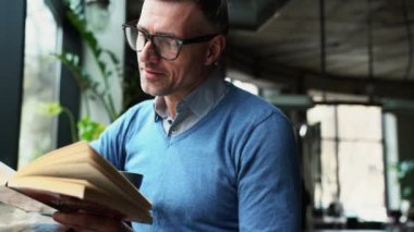 Gülümseyen gri saçlı adam kitap okuyor ve kafede otururken kahve içiyor.