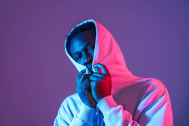 Siyah kapüşonlu genç adam poz veriyor ve mor duvardan izole edilmiş kameraya bakıyor.