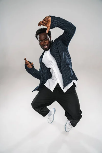 Young black man wearing jacket dancing and looking at camera isolated over white wall