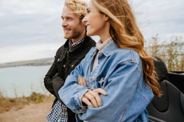 Beyaz, orta yaşlı bir çift, yolculuk sırasında arabasının yanında durup gülümsüyor.