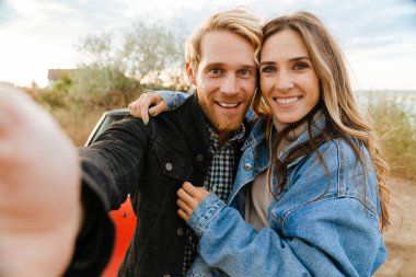 White mid couple making fun while taking selfie photo by car during trip