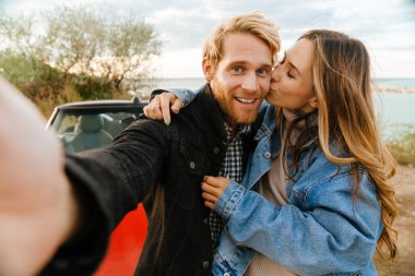 White mid couple smiling and kissing while taking selfie photo by car during trip