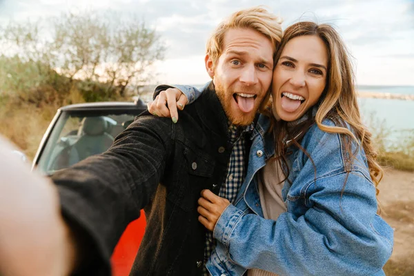 White mid couple making fun while taking selfie photo by car during trip