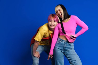 Asian teenagers with multicolored hair gesturing and laughing together isolated over blue wall