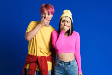 Asian shocked teenagers looking at camera and covering their mouths isolated over blue wall