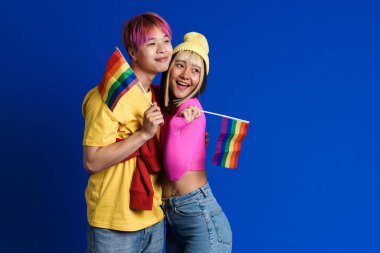 Asyalı gençler gülüp kucaklaşırken mavi duvarda izole edilmiş gökkuşağı bayraklarıyla poz veriyorlar.