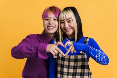 Asian couple smiling while showing heart gesture together isolated over yellow wall