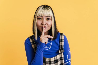 Sarı duvardan izole edilmiş kameraya sessiz jest yapan, piercingli bir Asyalı kız.