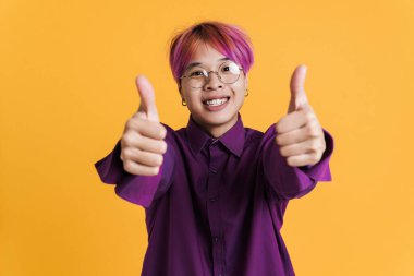 Gözlüklü Asyalı çocuk sarı duvarın üzerinde baş parmağını gösterirken gülüyor.