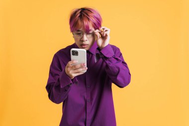 Sarı duvardan izole edilmiş gözlüklü Asyalı çocuk poz veriyor ve cep telefonu kullanıyor.