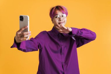 Gözlüklü Asyalı çocuk sarı duvarda izole edilmiş cep telefonuyla fotoğraf çekiyor.