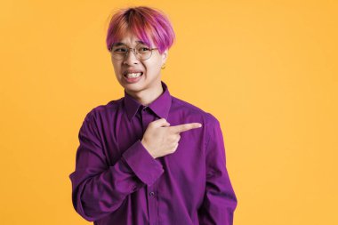 Asyalı hoşnutsuz çocuk yüzünü buruşturuyor ve sarı duvarın üzerinde soyutlanmış bir şekilde işaret ediyor.