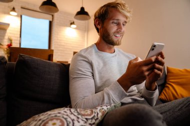 Kızıl saçlı adam evdeki koltukta otururken cep telefonu kullanıyor.