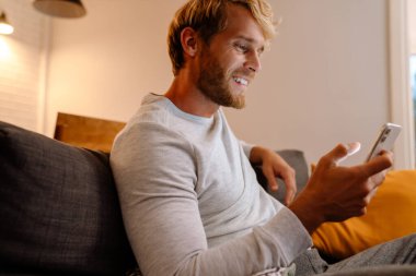 Kızıl saçlı adam evdeki koltukta otururken cep telefonu kullanıyor.