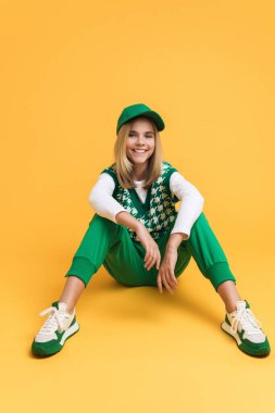 White girl wearing vest smiling and sitting on floor isolated over yellow background