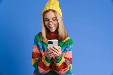 Gözlüklü beyaz kız gülümsüyor ve mavi arka planda cep telefonu kullanıyor.