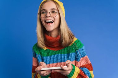 Gözlüklü beyaz kız mavi arka planda izole edilmiş cep telefonuyla çevrimiçi oyun oynuyor.