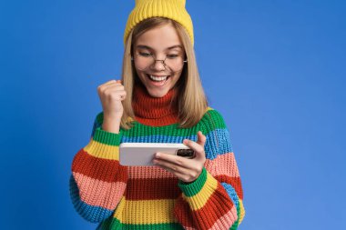 Gözlüklü beyaz kız mavi arka planda izole edilmiş cep telefonuyla çevrimiçi oyun oynuyor.