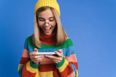 Gözlüklü beyaz kız mavi arka planda izole edilmiş cep telefonuyla çevrimiçi oyun oynuyor.