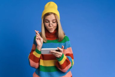 Gözlüklü beyaz kız mavi arka planda izole edilmiş cep telefonuyla çevrimiçi oyun oynuyor.