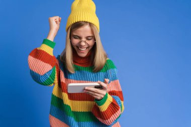 Gözlüklü beyaz kız mavi arka planda izole edilmiş cep telefonuyla çevrimiçi oyun oynuyor.