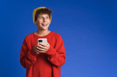 Şapka takan, gülümseyen ve mavi arka planda cep telefonu kullanan beyaz çocuk.