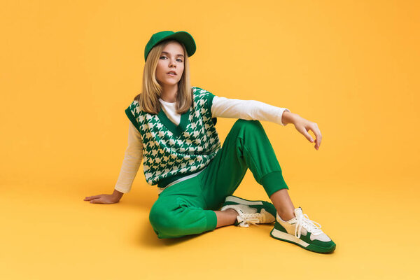 White girl wearing vest looking at camera and sitting on floor isolated over yellow background
