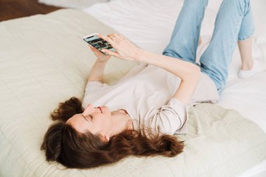 Beyaz kadın evde yatarken gülümsüyor ve cep telefonu kullanıyor.