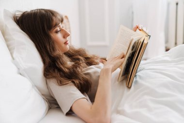 White young woman reading book while lying in bed at home