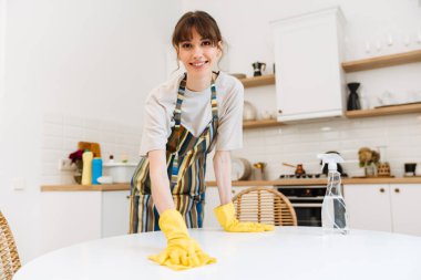 White young woman in gloves smiling while cleaning table at home