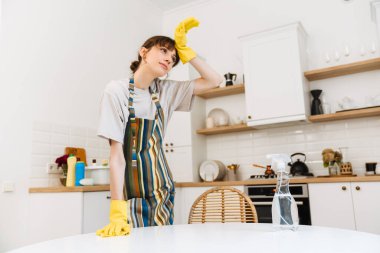 White young woman in gloves wiping her forehead while cleaning table at home
