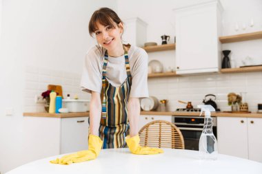 White young woman in gloves smiling while cleaning table at home