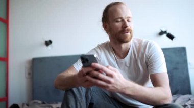 Cheerful bearded man texting by phone and looking to the side while sitting on the bed at home