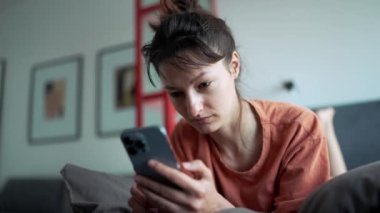 Serious brunette woman typing on phone while lying on the bed at home