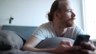 Pensive happy bearded man looking to the side and holding phone while lying on the bed at home