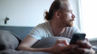 Thinking happy bearded man looking to the side and at the camera while lying on the bed at home