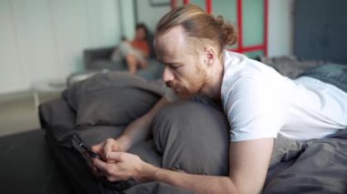Serious bearded man texting on mobile while lying on the bed at home